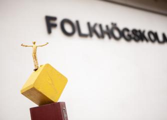 En skultpur med utsträckta armar framför en vägg med texten folkhögskola